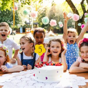 Seven kids celebrate a birthday at a picnic table outdoors on a sunny day with a Nostra Proin cake showing the number five, surrounded by flowers and paper pom-poms.