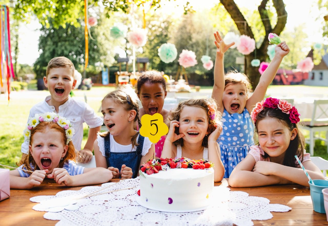 Seven kids celebrate a birthday at a picnic table outdoors on a sunny day with a Nostra Proin cake showing the number five, surrounded by flowers and paper pom-poms.