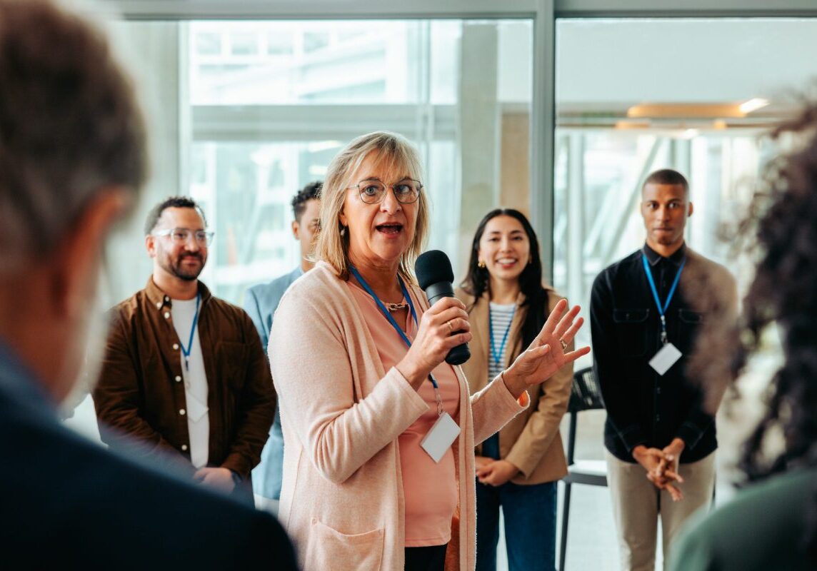 A woman speaks into a microphone at an indoor event. She stands in front of four people who are listening attentively.