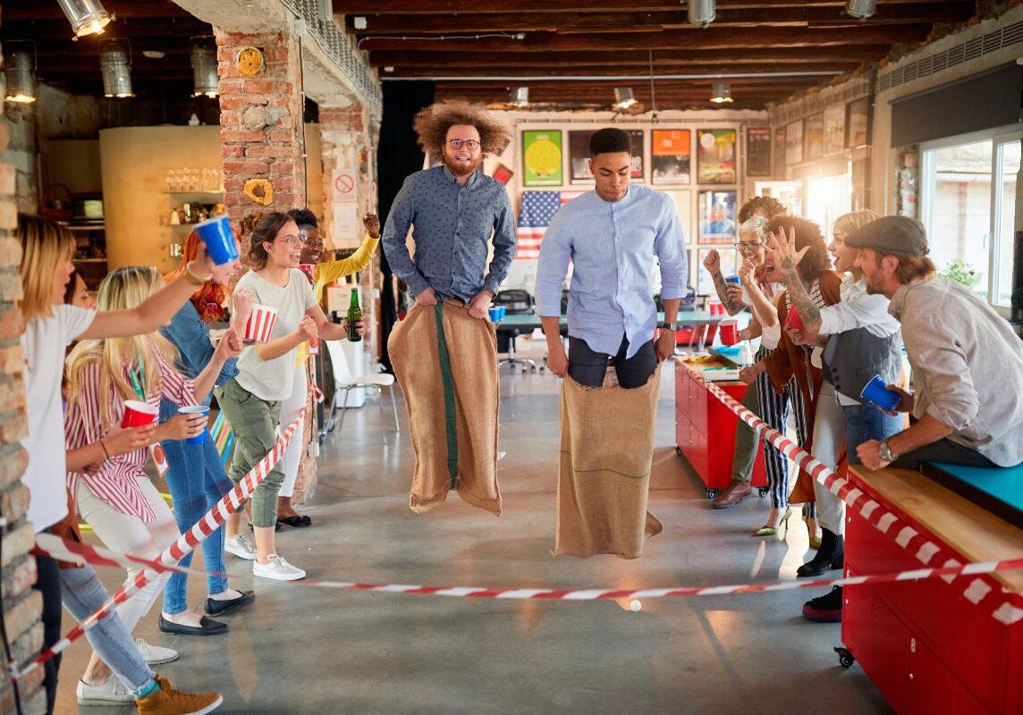 Two men compete in a sack race indoors, cheered on by a group of people. The room has exposed brick walls and colorful decor.